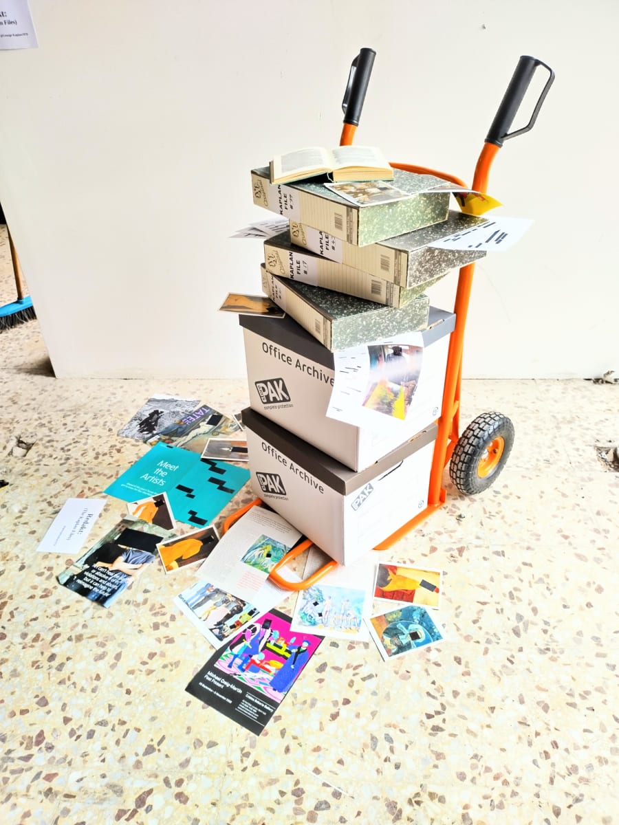 A sack barrow supporting archive boxes, box files and strewn with redacted books, art ephemera including artist statements and art images.
