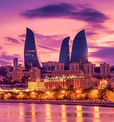 Baku skyline at sunset with illuminated Flame Towers reflected in Caspian Sea, historic buildings along waterfront