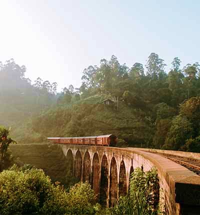 Red train crossing a historic arched bridge over a misty valley surrounded by lush green forested hills in Kandy, Sri Lanka