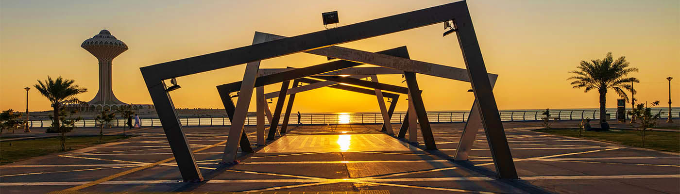 Sunset view through modern black metal structure at Dammam waterfront with water tower and palm trees in background