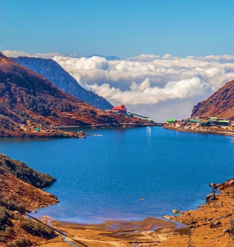 Scenic mountain lake surrounded by rocky peaks with a red-roofed building and settlement on the hillside under blue sky with 
