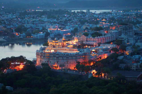 Udaipur city palace illuminated at dusk overlooking Lake Pichola with cityscape and mountains in background