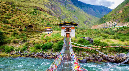 Traditional Bhutanese tower temple on wooden bridge over turquoise river in Paro valley surrounded by green mountains