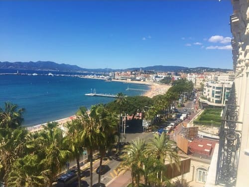 Aerial view of Cannes coastline with palm-lined promenade, sandy beach, turquoise Mediterranean Sea, and mountain backdrop