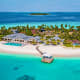 Aerial view of a tropical resort with white buildings, swimming pool, and wooden dock extending into turquoise lagoon with di