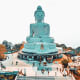 Giant turquoise Buddha statue seated on elevated platform with visitors and stairs at base in Phuket, Thailand