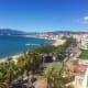 Aerial view of Cannes coastline with palm trees, Mediterranean Sea, sandy beach, and mountains in the distance under clear bl