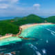 Aerial view of Coral Island with turquoise waters, sandy beach, green forested hills, and anchored boats