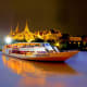 Tourist dinner cruise boat illuminated at night on Chao Phraya River with Golden Palace temple complex glowing in background,