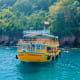 Bright yellow tour boat on turquoise water with lush green forested cliffs and red flag in background