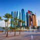 Abu Dhabi city skyline with modern skyscrapers, UAE flag, palm trees, and fountain in public plaza
