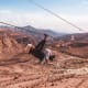 Person ziplining over desert mountains with red rock formations and winding roads in the valley below