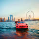 Red sports car floating in water with Dubai skyline and Ferris wheel at sunset