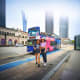 Two women walking past a colorful tour bus on a Kuala Lumpur street with modern skyscrapers and historic architecture in the 