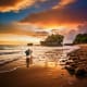Couple wading in shallow water at Tanah Lot temple during golden sunset with rocky coastline and dramatic orange sky