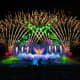 Grand stage with illuminated wings design, colorful light beams, and pyrotechnics during Wings of Time night performance