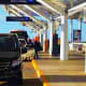 Black Mercedes vehicle at covered airport drop-off area with yellow curb marking and passengers walking in background