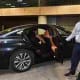 Man in business attire standing next to a black sedan with open door at indoor parking facility, Riyadh Airport