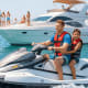 Man and child riding jet ski in front of luxury yacht with passengers, coastal city skyline in background