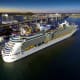 Aerial view of a large white cruise ship docked at a port terminal with blue water and coastal town buildings in background