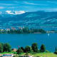Alpine lake view with snow-capped mountains, lakeside town with church, sailboat on turquoise water, and green meadows in for