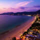 Aerial view of Pattaya beach at dusk with illuminated beachfront hotels, sandy shore, and mountains under purple twilight sky