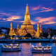 Wat Arun temple illuminated at dusk on the Chao Phraya River in Bangkok, Thailand, with traditional long-tail boats in foregr