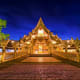 Illuminated golden Thai Buddhist temple with ornate roof and spires at twilight, wooden bridge and courtyard entrance