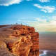 Visitors standing on rocky cliff edge overlooking vast desert landscape under blue sky with white clouds