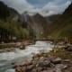 Mountain valley with rushing river, pine forests, and snow-capped peaks under cloudy sky in Kashmir