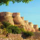 Ancient sandstone fort with crenellated walls perched on rocky hillside, surrounded by green vegetation under clear blue sky