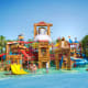 Colorful water park with large wooden-themed play structures, slides, and a massive bucket dumping water, surrounded by child