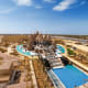 Aerial view of a large ornate temple with multiple spires surrounded by turquoise swimming pools and sandy desert landscape i