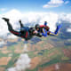 Three skydivers in formation freefalling above patchwork farmland with white clouds and blue sky
