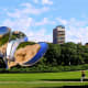 Large reflective metal flower sculpture in urban park with apartment buildings and green trees under blue sky