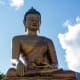 Seated bronze Buddha statue holding a scepter against blue sky with white clouds and forested mountains in background, Bhutan