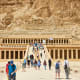Tourists walking toward the Temple of Hatshepsut in Egypt, with massive columned facade carved into limestone cliffs