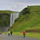 Tourists walking toward Skógafoss waterfall in Iceland, with cascading water falling from moss-covered cliffs surrounded by g