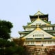 Osaka Castle with distinctive tiered roofs featuring green glazed tiles and gold ornaments, surrounded by palm trees against 