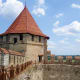Medieval stone fortress with conical red-tiled tower, wooden walkway, and green countryside landscape under blue sky in Moldo