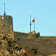 Ancient stone fortress with crenellated walls and towers overlooking rocky terrain under clear blue sky in Oman