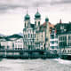 Historic Lucerne cityscape with white church featuring green onion domes reflected in the Reuss River, mountains in backgroun