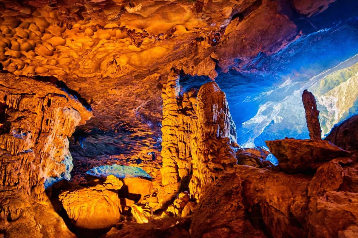 Underground cave chamber with golden limestone formations and blue lighting, stalactites hanging from ceiling, and natural sk