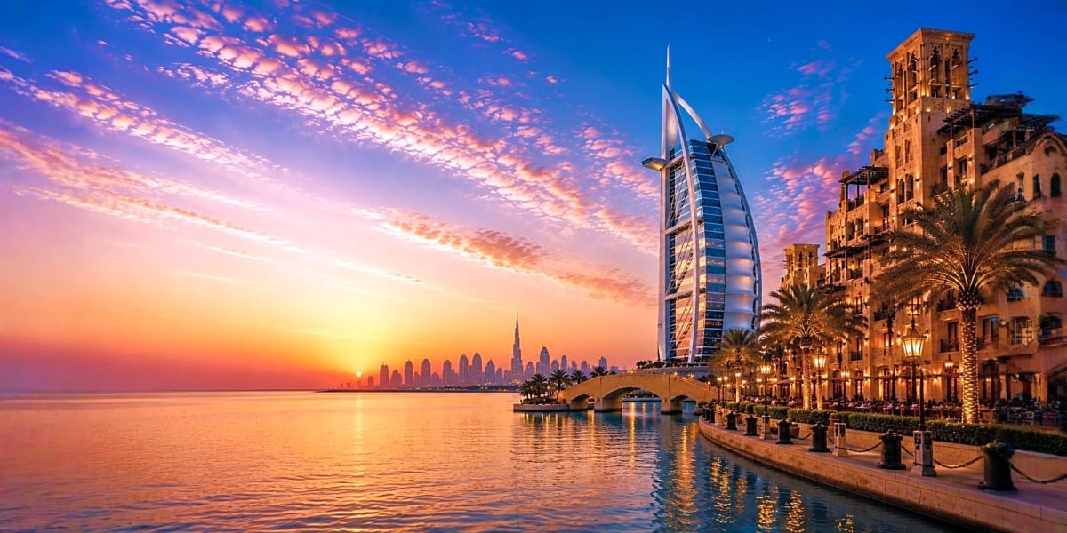 Dubai waterfront at sunset with Burj Al Arab hotel, palm trees, and illuminated buildings reflecting in calm Arabian Gulf wat