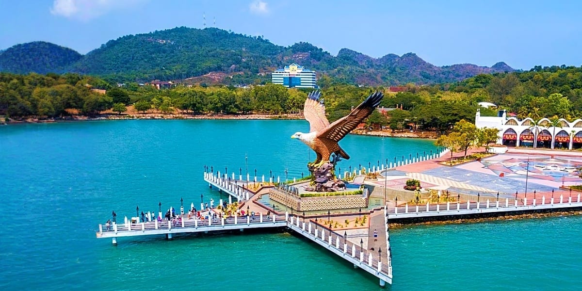 Eagle statue and white pier at Langkawi waterfront with turquoise sea, forested mountains and resort buildings in background