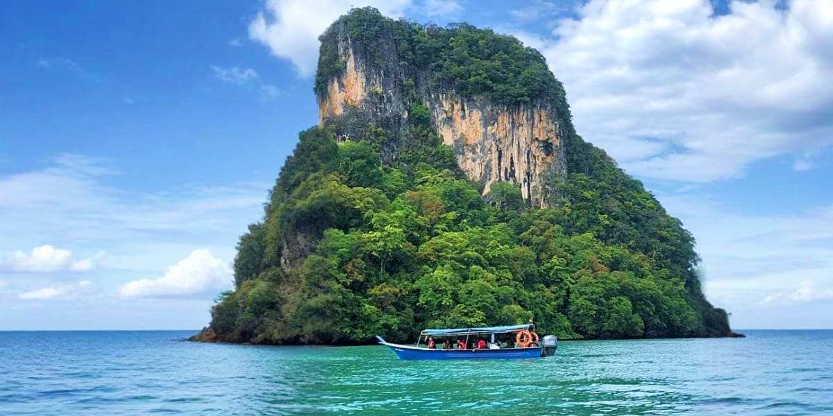 Limestone cliff island with dense jungle vegetation and turquoise water, blue longtail boat with tourists anchored nearby