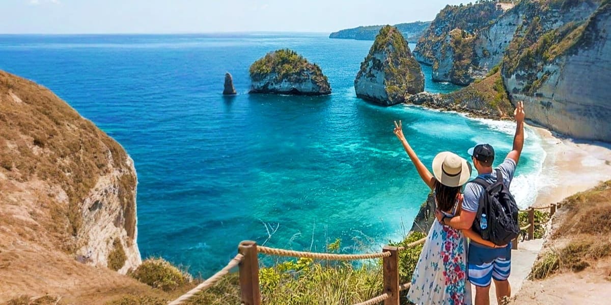 Couple with raised arms at clifftop viewpoint overlooking turquoise water, rock formations, and sandy beach in Bali