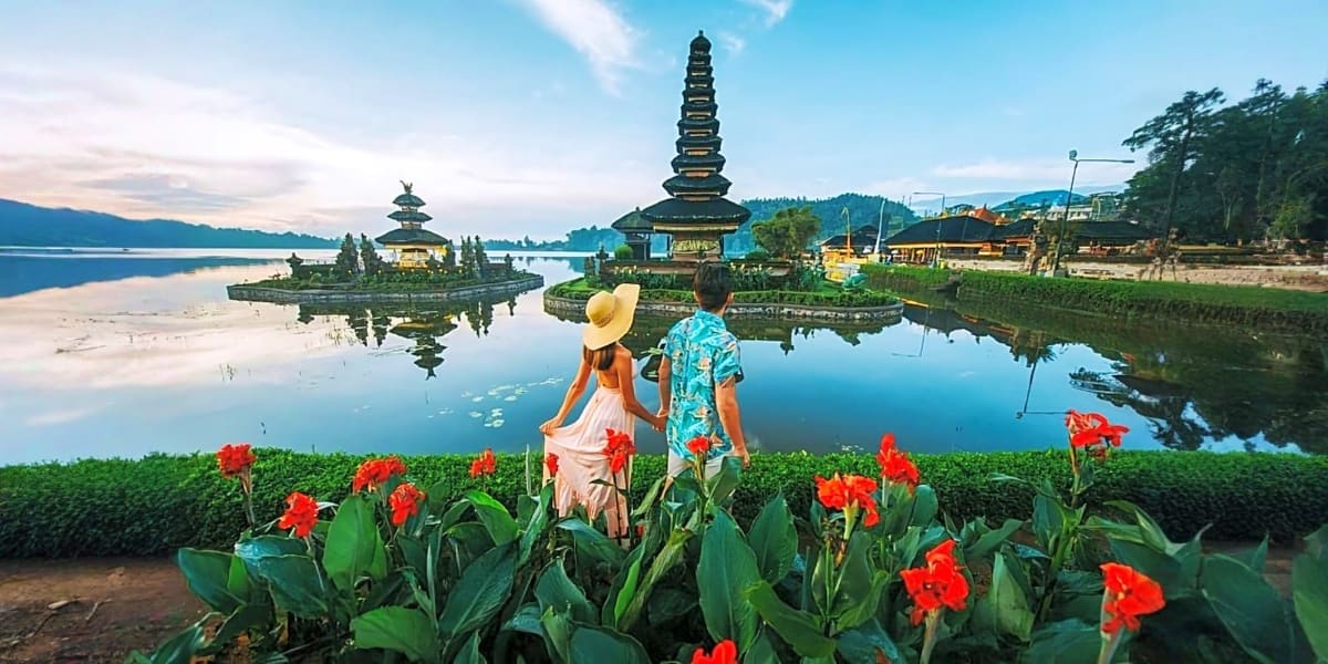 Couple viewing Pura Ulun Danu Bratan temple reflected in lake with red flowers in foreground, Bali Indonesia