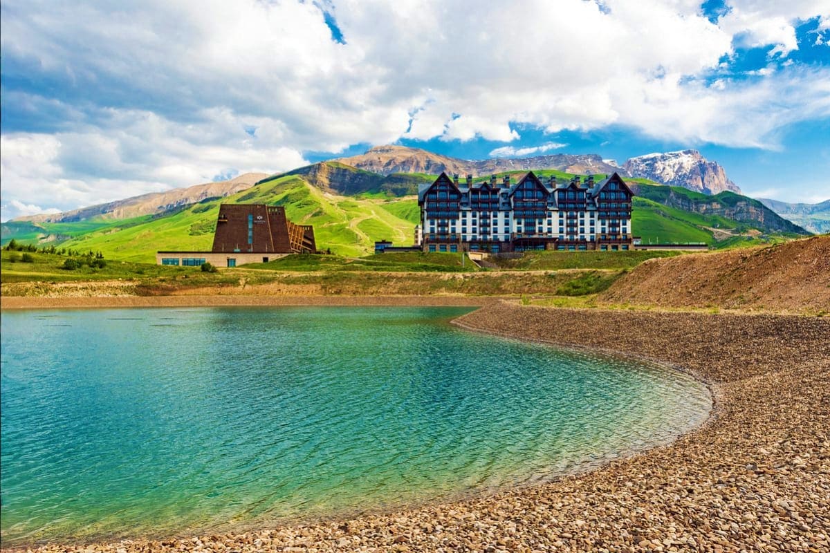 Alpine resort hotel on turquoise lake shore with green mountains and snow-capped peaks in background, Shahdag region Azerbaij