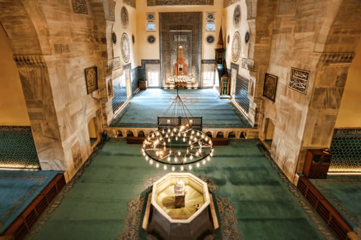 Interior of historic Ottoman mosque in Bursa with ornate chandeliers, green prayer carpets, and decorated stone walls with ca